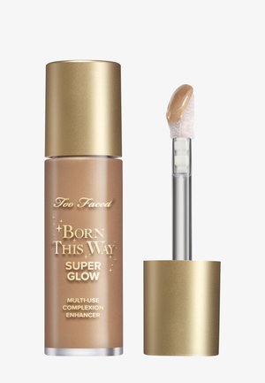 Too Faced Born This Way Super Glow enhanceerder, verpakt in een gouden dop met een matbruine fles en een transparante applicator met productpunt.