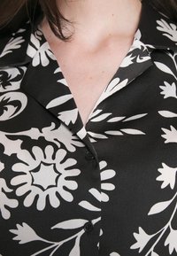 Chemise boutonnée à fleurs noire et blanche avec un col à revers, présentant de grands motifs floraux contrastés et une texture de tissu légère et lisse.