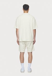 Uomo in piedi di spalle, con una camicia corta di tessuto color crema, pantaloni abbinati, calze bianche e sandali senza lacci su uno sfondo bianco.