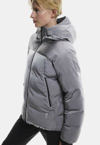 Jasnoszara kurtka puchowa z kapturem, z błyszczącym wykończeniem, zamek błyskawiczny z przodu, dwa boczne kieszenie i szczegół z logo z przodu.