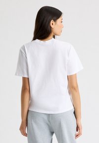Camiseta de algodón blanca con mangas cortas, un escote redondo y un corte holgado, vista desde atrás, combinada con pantalones de chándal grises.