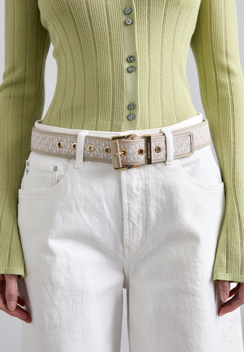 Cintura di crema con hardware dorato e banda decorata, indossata sopra jeans bianchi e una maglietta a coste verde chiaro con bottoni e maniche lunghe.
