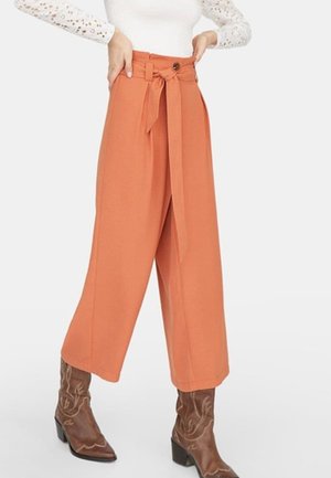 Vrouw die hooggetailleerde, wijd uitlopende oranje broek draagt met een geknoopte riem, gecombineerd met bruine geborduurde cowboylaarzen en een wit kanten top met lange mouwen.
