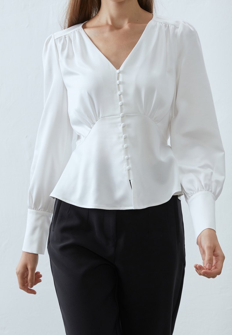 Femme portant un chemisier blanc à manches longues avec des boutons sur le devant et un pantalon noir, sur un fond clair uni.
