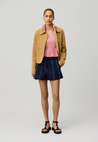 Giacca in suede tan sopra una polo a righe rosse e bianche, completata da pantaloni corti blu navy con cuciture bianche e sandali neri con cinturini.