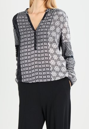 Femme portant un chemisier à manches longues avec des motifs géométriques et floraux noirs et blancs, associé à un pantalon noir, mains dans les poches.