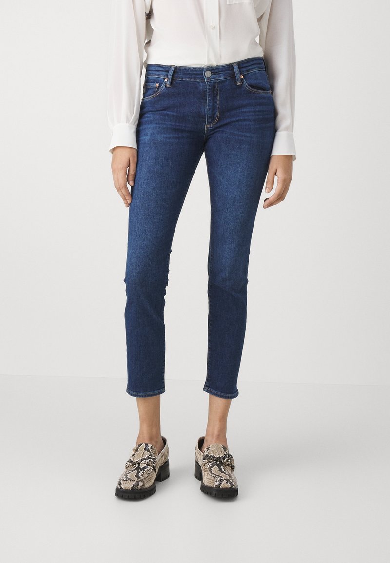 Jeans skinny azul oscuro con un corte ajustado, bolsillos frontales y un ligero deshilachado en el dobladillo, combinados con zapatos de tac ón grueso y estampados.