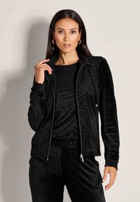 Vrouw met lang, donker haar die gouden oorbellen draagt, een zwart fluwelen jack met luipaardpatroon en bijpassende broeken.