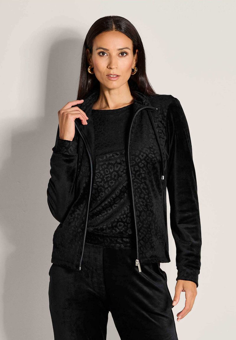 Vrouw met lang, donker haar die gouden oorbellen draagt, een zwart fluwelen jack met luipaardpatroon en bijpassende broeken.