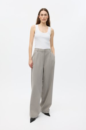 Pantaloni grigio chiaro a gamba larga con pieghe, vita alta, tasche laterali e texture liscia, abbinati a un top bianco aderente e scarpe nere con punta affilata.