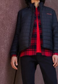 Padded black jacket z wysokim kołnierzem, z czerwonym logo "HUGO", noszony na czerwono-czarnej koszuli w kratkę, zestawiony z ciemnymi dżinsami, ręce spoczywające po bokach.