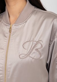 Veste bomber en satin taupe avec les initiales élégantes "LB" brodées sur la poitrine et une fermeture éclair dorée.