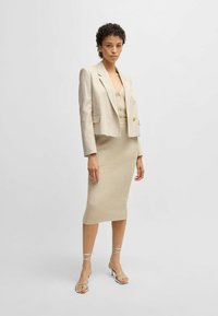 Ecru blazer met een cropped ontwerp over een ribgebreide ecru jurk. De outfit heeft knopen en sandalen met strappy en hoge hakken. Gladde textuur.