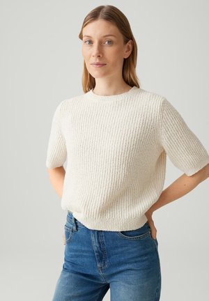 Frau mit hellbraunen Haaren, die einen strukturierten weißen Kurzarmpullover und blaue Jeans trägt und mit einer Hand hinter dem Rücken steht.