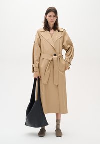 Beige trench coat with a belted waist, large collar, and button accents. The model holds a black leather tote and wears brown slip-on shoes.