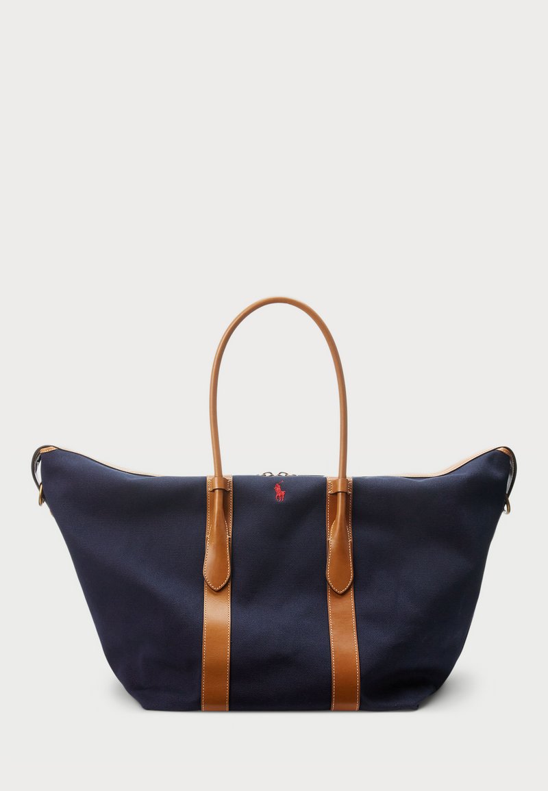 Saco duffel de lona azul-marinho com alças de couro bege, fecho com zíper e pequeno detalhe em logo vermelho na frente, apresentando um design amplo e espaçoso.