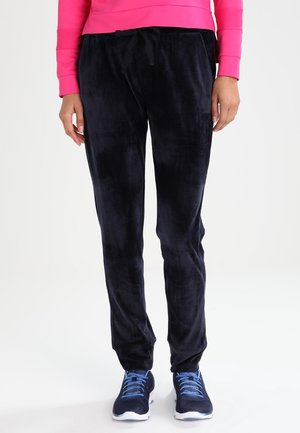 Persona con pantalones de velour negros con cordón, zapatillas azules con cordones azul claro, y una camiseta de manga larga rosa brillante, de pie sobre un fondo blanco.