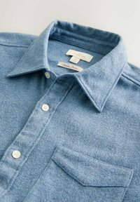 Šviesiai mėlynas mygtukinis marškinis su smailiu apykakle, krūtinės kišene ir baltais mygtukais, pagamintas iš minkštos tekstūruotos medžiagos.