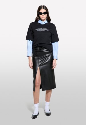 T-shirt noir avec un design graphique, superposé à une chemise à col rayée bleue. Jupe en cuir noir avec une fente à l'avant, associée à des chaussettes blanches et des chaussures noires.