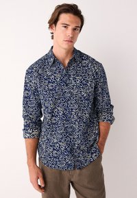 Tumesinine nööbitav särk kreemika lillemustriga ja üles keritud varrukatega, kombineeritud helepruunide pükstega. Kangas tundub sile ja kerge.