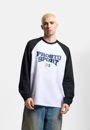 Młoda osoba z krótkimi blond włosami, ubrana w białą i czarną koszulkę z długim rękawem z napisem "PROSTO SPORT" oraz niebieskie dżinsy.