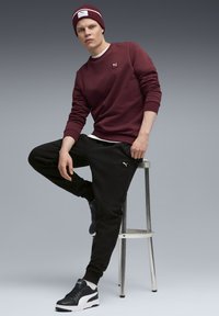 Bordeauxfarbenes Sweatshirt mit kleinem Logo, schwarze Sweatpants und schwarze Turnschuhe. Modell sitzt auf einem Hocker vor einem verlaufenden grauen Hintergrund.