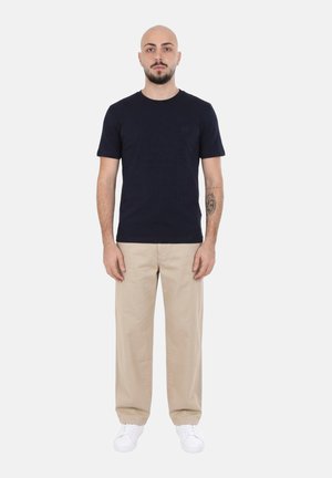 Uomo calvo con barba che indossa una maglietta nera, pantaloni beige e scarpe da ginnastica bianche, in piedi dritto su uno sfondo bianco semplice.