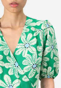 Groene jurk met een bloemenpatroon met grote witte bladeren en blauwe accenten. De stof lijkt lichtgewicht met puffed mouwen en een V-hals.