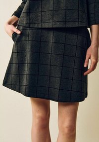 Kvinna klädd i mörkgrå kjol och topp med svart rutmönster, ena handen i fickan och den andra avslappnad längs sidan.