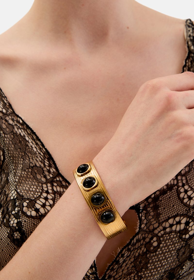 Złota bransoletka mankietowa o żebrowanej fakturze z czterema owalnymi czarnymi kamieniami. Noszona na nadgarstku na tle czarnej koronki.
