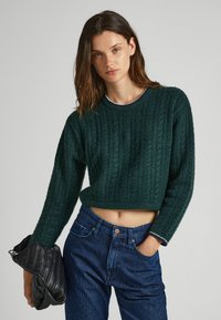 Maglione corto di un verde scuro, con una texture e un design a coste, dotato di un collo rotondo. Abbinato a jeans di denim a strisce blu scuro e una borsa nera.