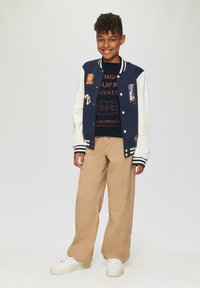 Marineblau und weiße Collegejacke mit Aufnähern, über einem schwarzen T-Shirt mit Grafik, kombiniert mit beigen weiten Hosen und weißen Sneakers.