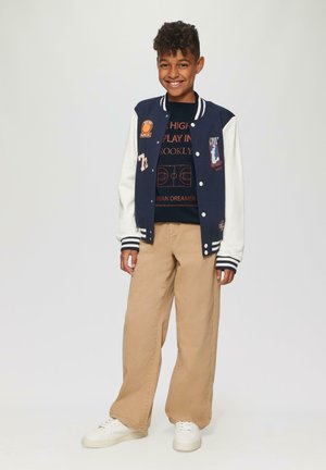Marineblauwe en witte varsity jacket met patches, gedragen over een zwart graphic t-shirt, gecombineerd met beige wijde broek en witte sneakers.