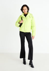 Neon green puffer jacket with a stand collar, snap closures, and pockets, paired with black pants and heeled ankle boots.