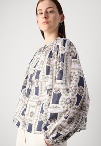 Blusa bianca e blu navy con motivi a palloncino, caratterizzata da disegni circolari e geometrici, tessuto strutturato e scollatura arrotondata.