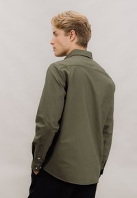 Oliven grønn skjorte med knapper foran, lange ermer, en rett fasong og en jevn stofftekstur. Har standard mansjetter og en ryggklaff.