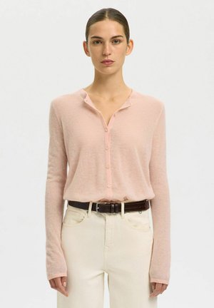 Kvinde iført en lyserød knap-cardigan stoppet ned i cremefarvede højtaljede bukser med et mørkebrunt bælte, stående foran en ensfarvet baggrund.