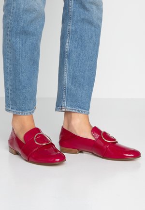 Des pieds chaussés de mocassins rouges brillants avec une grande boucle dorée, associés à un jean en denim bleu clair sur un fond uni clair.