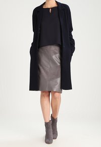 Marineblauer langer Mantel über einem dunklen Oberteil, kombiniert mit einem grauen Kunstledrrock mit Chevron-Panels und grauen Stiefeletten mit seitlichem Reißverschluss.