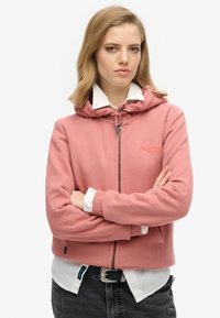 Sudadera con cremallera rosa y capucha con cordón, textura suave, logo bordado en el pecho y puños acanalados, llevada sobre una camisa blanca con cuello.
