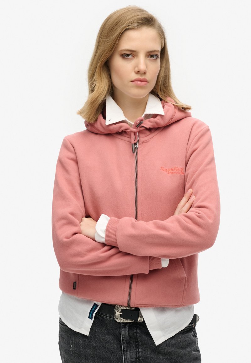 Sudadera con cremallera rosa y capucha con cordón, textura suave, logo bordado en el pecho y puños acanalados, llevada sobre una camisa blanca con cuello.