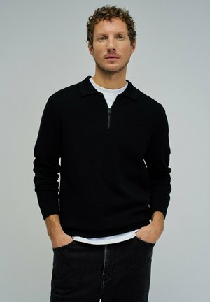 Hombre con cabello rizado, vistiendo un suéter negro con cierre sobre una camisa blanca, pantalones negros, de pie con las manos en los bolsillos, fondo neutro.