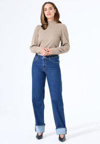 Beige geripptes Strickpullover mit Puffärmeln, kombiniert mit hochgeschnittenen blauen Jeans, die an den Knöcheln umgeschlagen sind. Schwarze spitze Absatzschuhe.