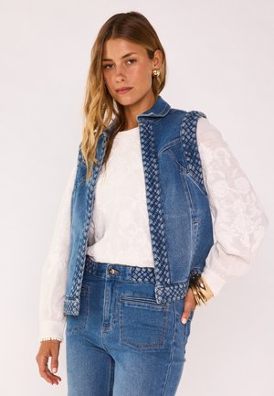 Femme portant un gilet en jean tressé et un jean avec un chemisier blanc brodé de motifs floraux, des boucles d'oreilles en or et plusieurs bracelets en or aux poignets.