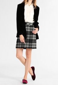 Blazer en velours noir, blouse blanche, jupe en laine à carreaux gris et noir, et mocassins rouge foncé. Le mannequin se tient avec une main sur la hanche.