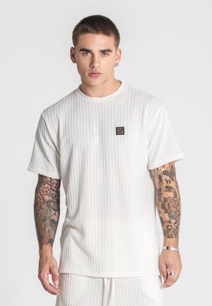 Witte T-shirt met korte mouwen en een gestructureerd ontwerp, ronde hals en een klein logo-embleem op de linkerborst. Bevat zichtbare tatoeages op de armen.