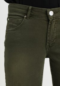 Vaqueros verde oliva hechos de denim duradero. Cuenta con cinco bolsillos, cremallera y acentos de botones y remaches metálicos plateados. Textura suave.
