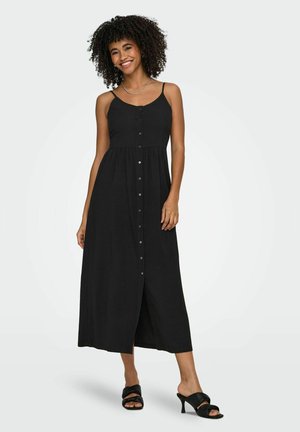 Femme aux cheveux bouclés souriante, portant une robe midi noire sans manches avec des boutons sur le devant et des sandales à talons noires, debout devant un fond uni.