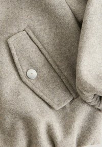 Jasnoszara wełniana kurtka o miękkiej fakturze. Wyposażona w dużą kieszeń z klapą oraz okrągły metalowy guzik. Tkanina wydaje się gruba i ciepła.