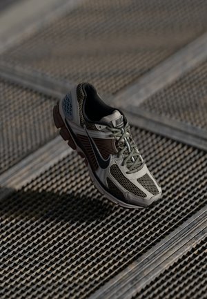 Ein einzelner grauer und schwarzer Nike Laufschuh mit Mesh-Design, der über einer strukturierten Metallgitterfläche schwebt.
